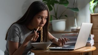 A woman sitting at a table, eating ramen and interacting with a laptop