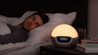 The image shows a man laying in bed with a Lumie Bodyclock Shine 300 glowing gently on a bedside table next to him