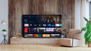 A Mitchell & Brown 65-inch 4K TV on a TV cabinet in front of a wood-panelled wall. On screen are rows of apps and content stills.