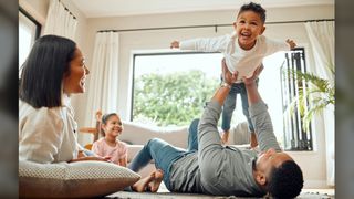 We see a mom, dad, daughter and son playing in a living room. The dad is lying on the floor and playing "airplane" with his son, whose arms are outstretched. 