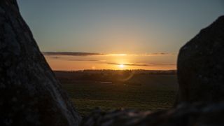 the sun is visible far in the background, above trees and amid clouds. two stones frame the sides of the picture