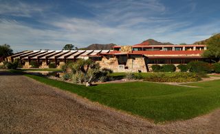 Taliesin West designed by Frank Lloyd Wright and built in 1937. A large house made from stones with a wooden roof, beautiful gardens in front of it and hills behind it.