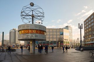 The World Clock in Berlin shows global times and has a large structure of the solar system on top, it is surrounded by people and buildings. 