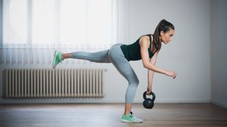 Woman at home on plain background performing a single-leg Romanian deadlift with left leg back, holding kettlebell in left hand, right arm extended