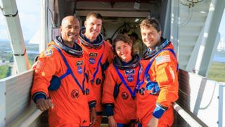 four astronauts in orange pumpkin suits in a row. they stand on a crew access arm that usually brings them to a rocket. behind is the view of the florida water