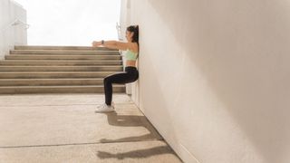 Woman performing a wall sit outdoors in the sunshine with both arms extended