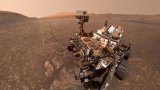 a large, metallic mars rover sits on the red dirt, with small hills in the background.