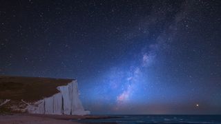 a large cliff structure on the left with white rock face and grass on top, with the milky way visible to the right over the sea.