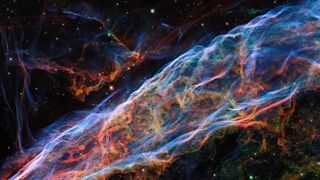 a wispy nebula colored red, blue, orange and pink against a black background speckled with stars. 