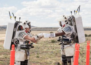 two people in skeleton-like spacesuits without the outer shell walk through the desert under a bright sun