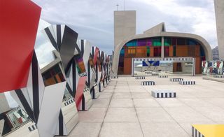 To the right, we see square-shaped structures set on the rooftop. Each structure has black & white lines on its side, and the top is in different colors - yellow, blue, and white. The structures are lined up to the reflective surface which gives us the impression of infinite rows of blocks. To the left, we see different colored panels that are connected in the middle by either protruding at a sharp angle or receding.