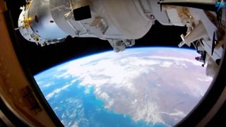 a white spacecraft is seen through the window of a space station, with earth in the background.