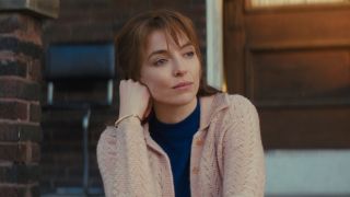 Jodie Comer sits on a stoop with a wistful smile on her face in The Bikeriders.