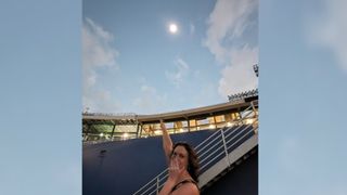 A woman with shoulder length brown wavy hair holds one hand over her mouth and her other hand points up at the sun during the April 8 total solar eclipse.