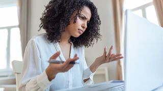 A woman sitting as a desk, looking at her laptop and expressing frustration with her face and hands