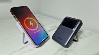 Two UGREEN Uno 15W Qi2 power banks are side by side on a marble effect surface, angled slightly toward each other. The power banks are gray and purple, the latter has an iPhone 12 Pro charging on it.