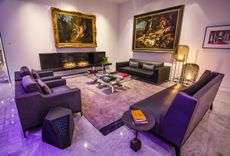 A lounge area with a purple leather sofa, purple fabric chairs, a black leather sofa, a fireplace, floor lamps, coffee tables, a glass table and large wall paintings.