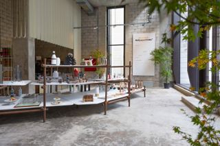 'The shop' at the House of Today. In the room, there is a large and wide shelf with different houseware and kitchenware products. Bottles, glasses, cake plates, etc are set on white top shelves.