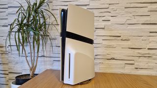 The PS5 Pro console on a wooden desk in front of a white brick background and next to a potted plant