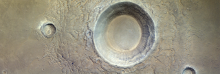 A high aerial view above a rocky, desolate landscape, with a deep, enormous crater scooped out from nearly half the image on the right. There is also a much smaller crater on the left.