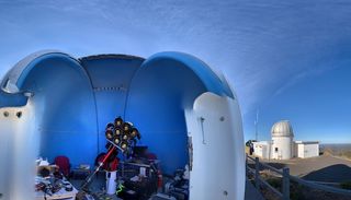 an open sided dome structure hovers above telescope equipment in the foreground. behind to the right, a domed silo with blue skies above.