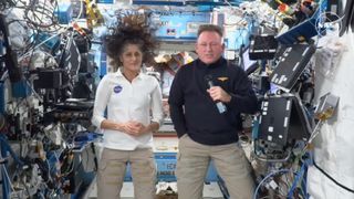 a woman in a white shirt and a man in a blue shirt stand inside a crowded space module