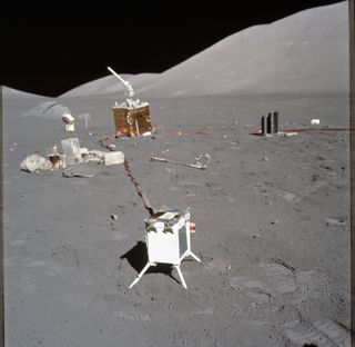 A photograph taken during Apollo 17 shows science experiments in the foreground and background with a debris pile including experiment wrappers and covers on the left.