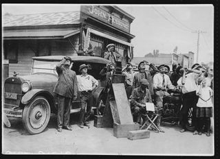 A crowd looks up to the sky, there is a telescope in the center and a car in the background. People of all ages are there for the event.