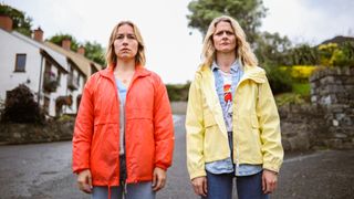 Sare (Sarah Goldberg) and Suze (Susan Stanley) stand next to each other wearing brightly coats in SisterS