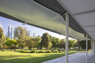 View from the white frame pavilion, looking out across the grassed lawn, surrounding trees and shrubs, high rise buildings in the distance against a blue sky