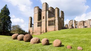 Eight stone catapult shots sit in the grass in front of a medieval castle. 