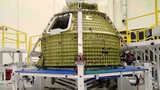 a partially completed conical gold space capsule sits on a metallic frame inside a white-walled building