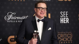 Brendan Fraser stands dressed in a black tuxedo, holding his Critic's Choice Award. 