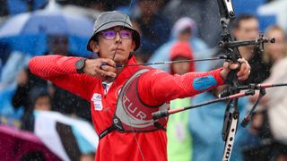 Olympic Archery champion Mete Gazoz of Turkiye aims his bow wearing a red shirt and grey bucket hat.