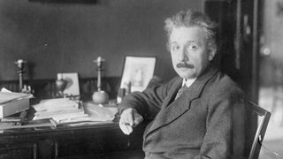 A black and white photograph of Albert Einstein wearing a suit and sitting at his desk