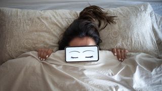 A woman lies in bed with a phone covering her eyes