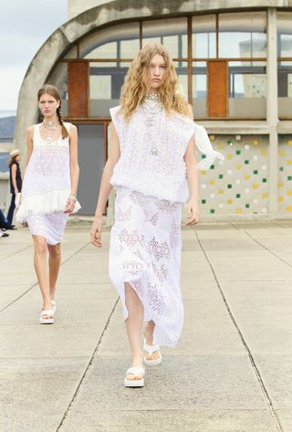 Models in white Chanel dresses walk in runway show on concrete rooftop