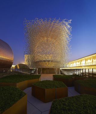 View of the UK Pavilion featuring a tall, aluminium mesh ’beehive’ structure and several patinated metal, geometric planters with low greenery in the evening