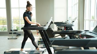 12-3-30 treadmill workout: Woman walking on a treadmill during workout in the gym with headphones around her neck