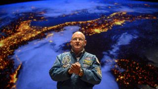 a man in a blue flight jacket stands in front of a map of Earth