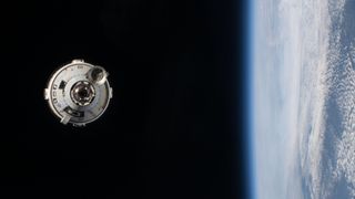 a white space capsule hangs in the blackness of space, with earth to the right of the frame