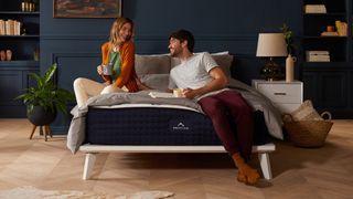 The best mattress in a box: DreamCloud Mattress on a wooden bedframe placed in a dark blue bedroom and with a couple laughing and sitting on the mattress