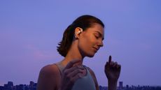 A woman wearing an athletic tanktop and the white Shokz OpenFit 2 wireless earbuds with her eyes closed, listening to the music, with a purple sky and a distant building skyline behind her.