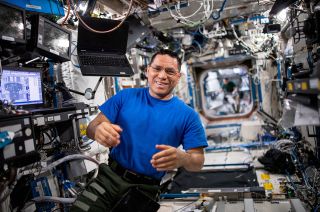 a smiling man in a blue shirt floats aboard the space station with computers and wires in the background.