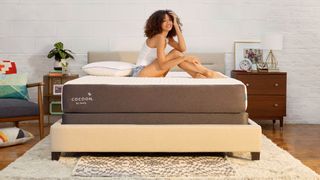 A woman with dark curly hair sits on the Cocoon by Sealy Chill mattress