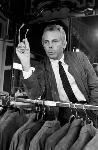 Giorgio Armani in a suit, holding a pair of glasses with his right hand over a rail rack of jackets