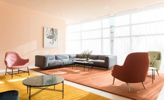 A sitting area with three chairs, a round table, a rectangular table, a yellow rug, an orange rug, a wall painting and a grey corner sofa next to a large window with white curtains.