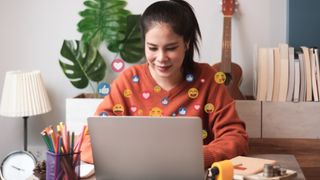 A person using a laptop with emoji symbols floating around the laptop, suggesting she is using emoji on her computer