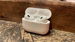 Apple AirPods Pro 2 in their charging case on a scratched wooden surface against a darker wooden wall.