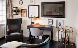 A white sitting room with a black leather chair, a couch, a desk, wall paintings and a standing lamp next to a window.